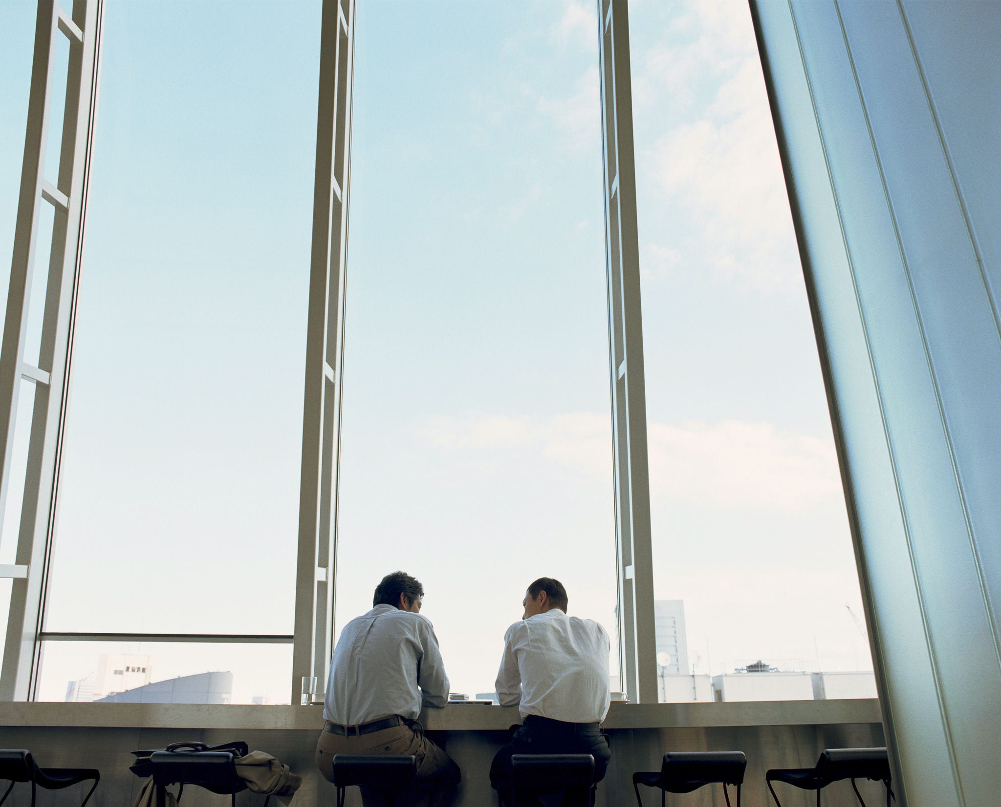 Two men in a high-rise office building sit in front of large open windows, overlooking the city below.